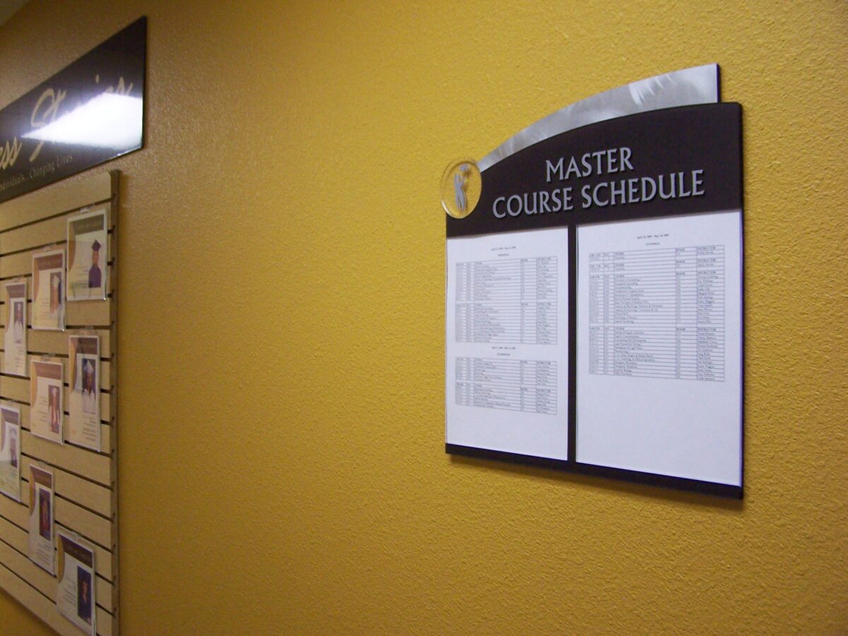 A wall-mounted display titled "Master Course Schedule" on a yellow wall. The schedule consists of two white sheets with text. Nearby on the left, a bulletin board contains various posted items, including photos and papers. The scene is well-lit.