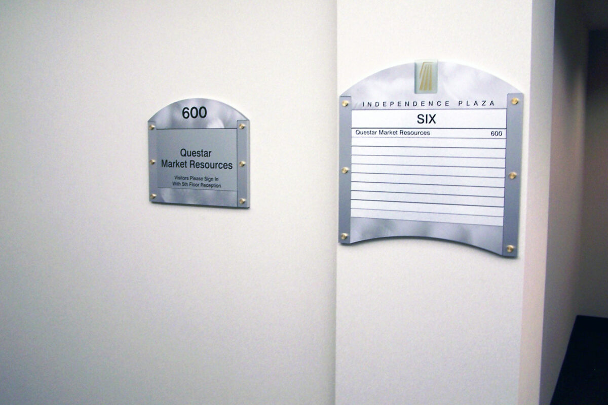 Wall-mounted signs for offices. One sign reads "600," with "Quarter Market Research" and additional details below. Another larger sign titled "SIX" has a list of individuals and office numbers. Both are mounted on a white wall next to an office cubicle.