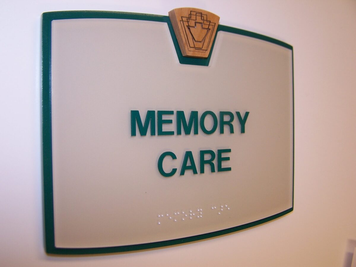 A frosted glass sign with green borders and a wooden emblem at the top, displaying the words "MEMORY CARE" in green uppercase letters. Below the text, there is braille writing. The sign is mounted on a white wall.