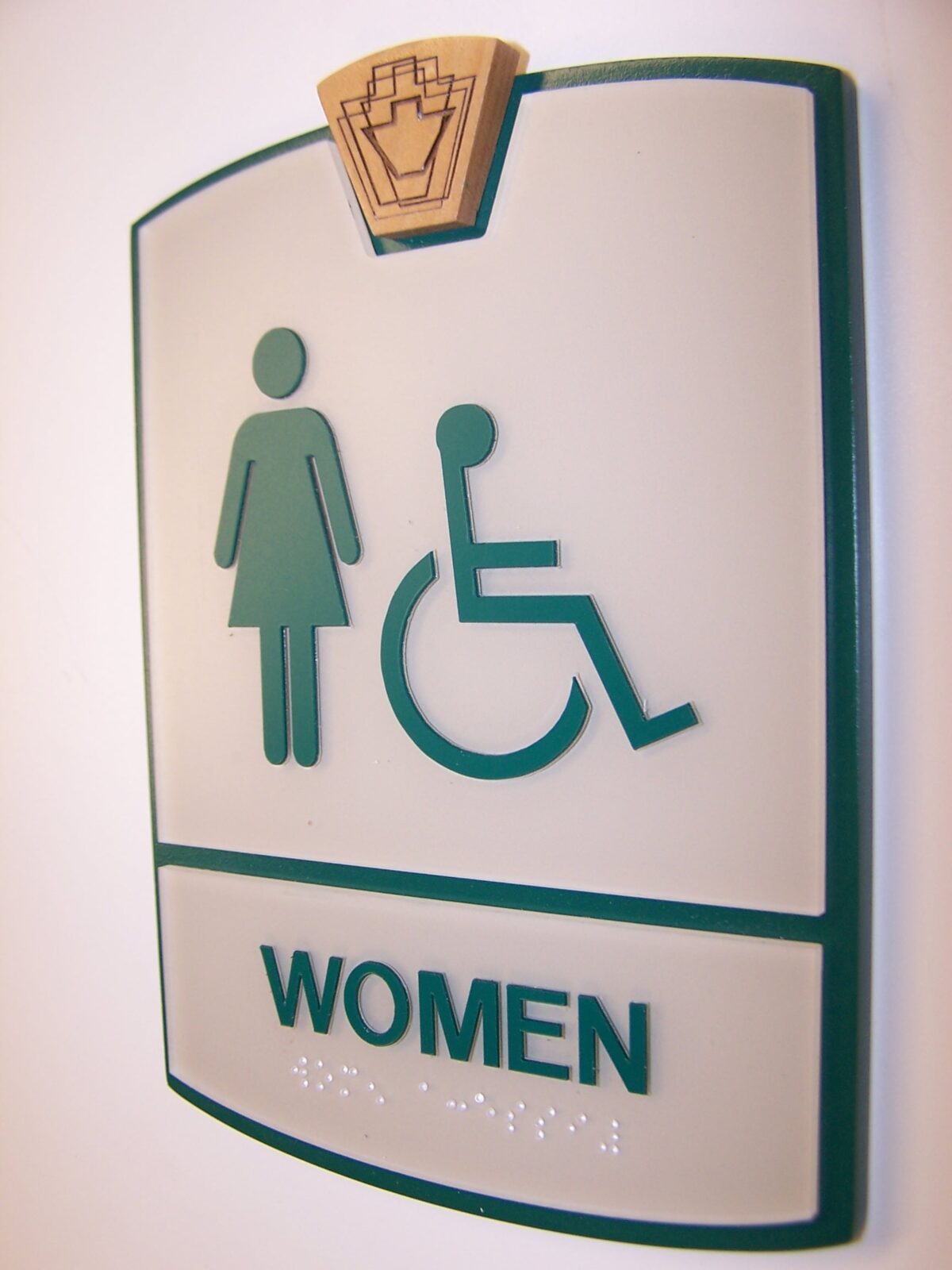 A restroom sign for women with a green border. The sign features symbols for a woman and a person in a wheelchair, indicating the restroom is accessible. The word "WOMEN" is written below the symbols in green text. Braille is included beneath the text.