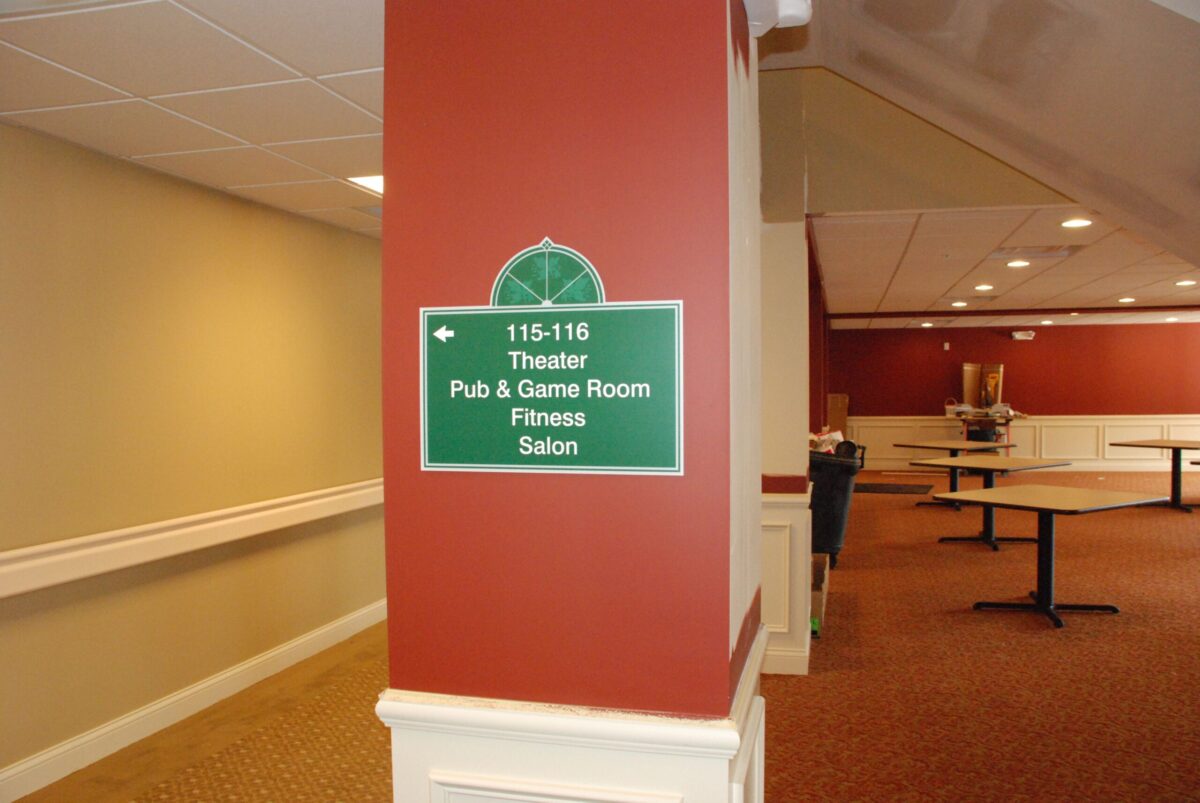 A green rectangular sign on a red column in a hallway. The sign provides directions to various facilities, including Theater, Pub & Game Room, Fitness, and Salon for rooms 115-116. Surrounding area includes beige walls, a white chair rail, and a carpeted floor.