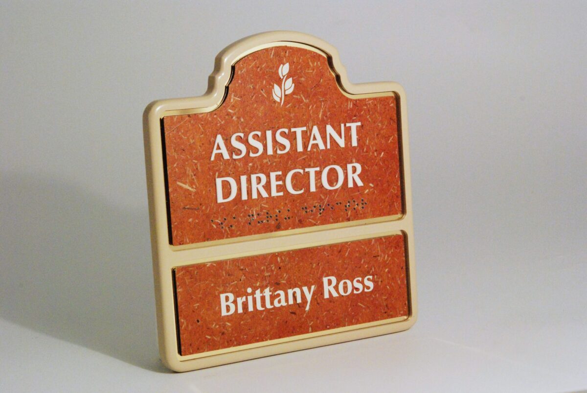 A sign with a red background and a beige border. The top part reads "ASSISTANT DIRECTOR" in white text, while the bottom part displays the name "Brittany Ross" in the same white text.