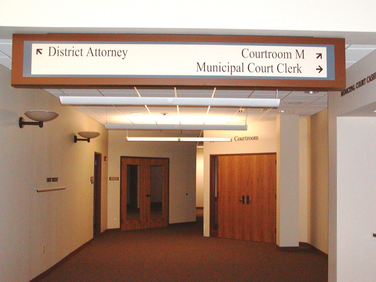 A hallway in a building with signs on the ceiling directing to the offices of the District Attorney, Municipal Court Clerk, and Courtroom M. The walls are cream-colored and several doors are visible along the corridor. Wall sconces provide lighting.