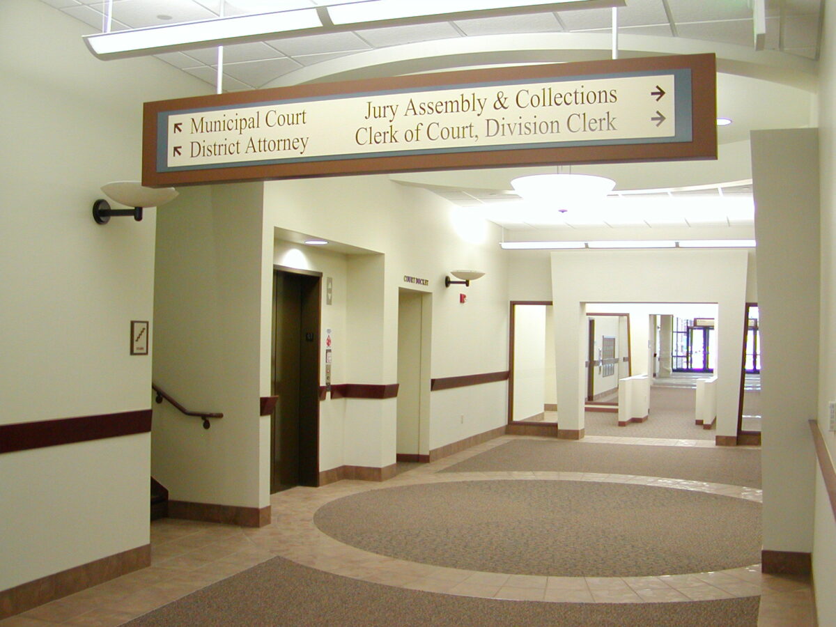 A hallway inside a courthouse with a signpost hanging from the ceiling. The sign directs to Municipal Court, District Attorney, Jury Assembly & Collections, and Clerk of Court, Division Clerk. The corridor features beige walls and carpeted floors.