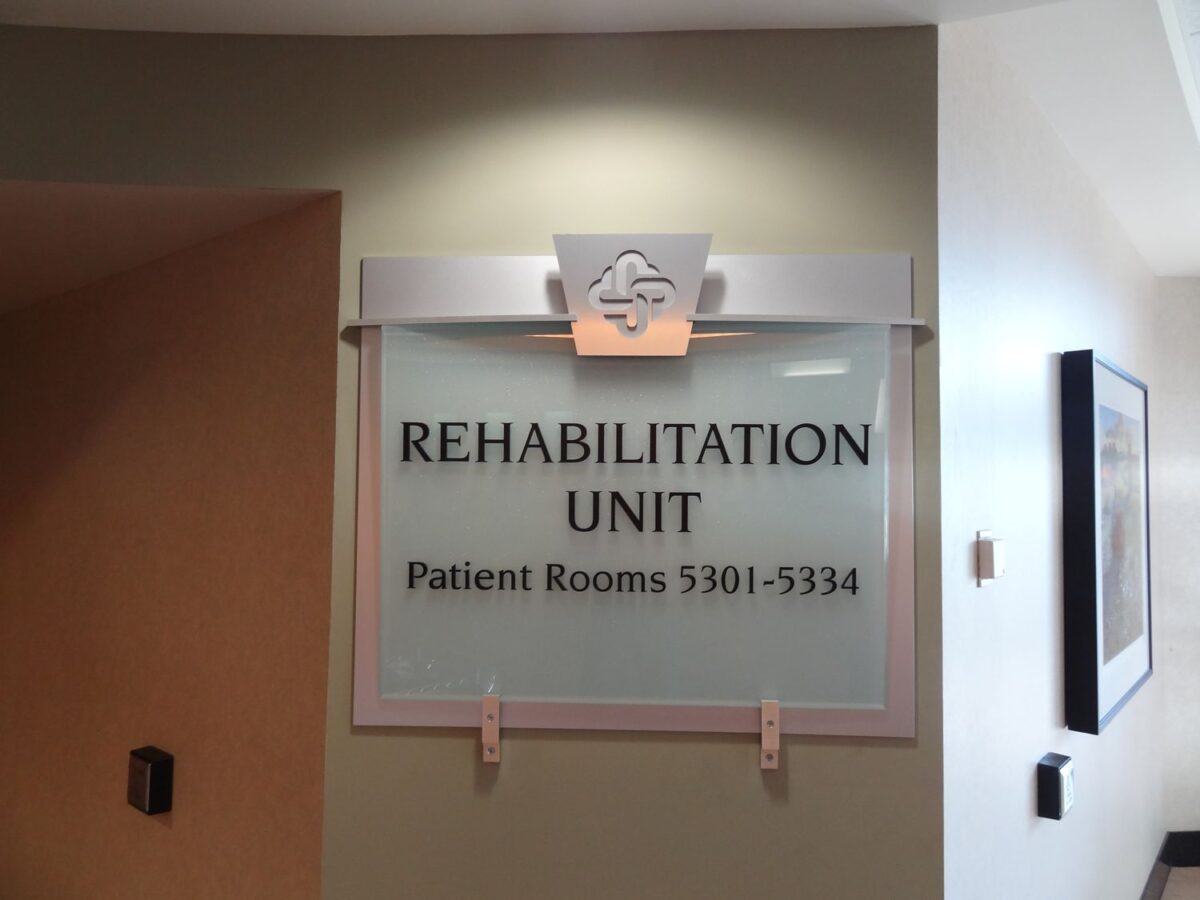 A sign mounted on a light-colored wall indicates the location of a Rehabilitation Unit in a medical facility. The sign includes room numbers for patient rooms, specifically 5301 to 5334. The design features a medical emblem above the text.