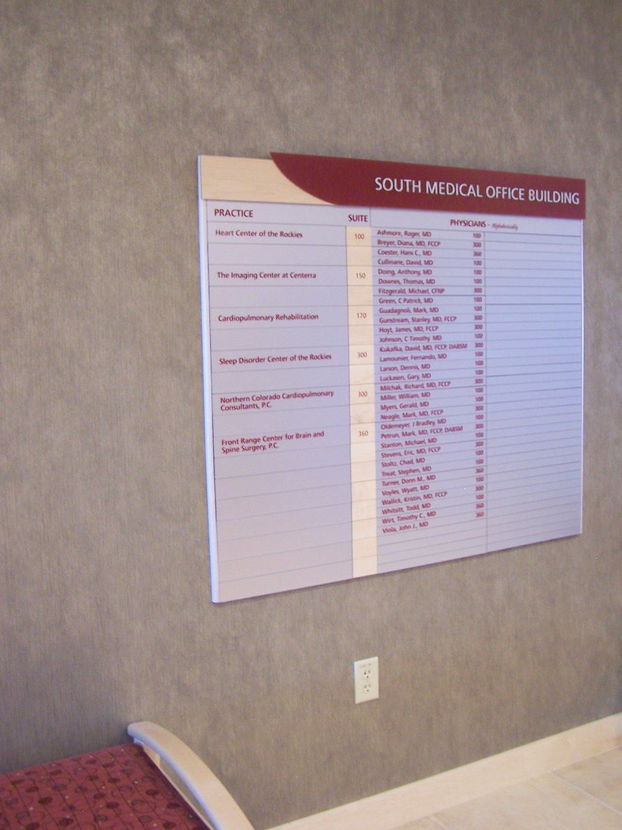Directory board on a gray wall in the South Medical Office Building. The board lists various office numbers, doctor names, and specialties. A small portion of a cushioned chair is visible at the bottom left corner of the image.