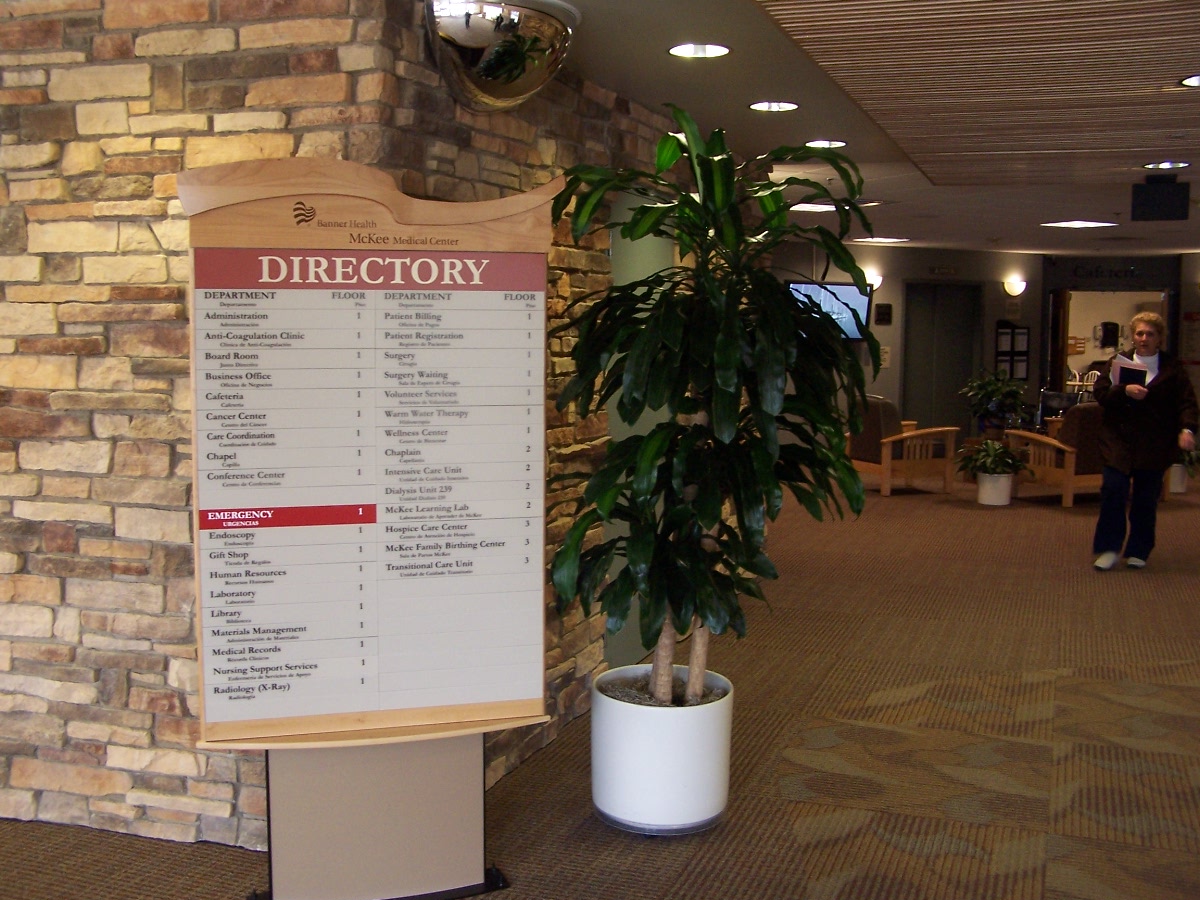 A directory sign is displayed in a hallway with a stone wall on the left. The sign lists different locations within the building. Adjacent to the sign is a large potted plant. The hallway leads to more rooms in the background, with a few people visible.