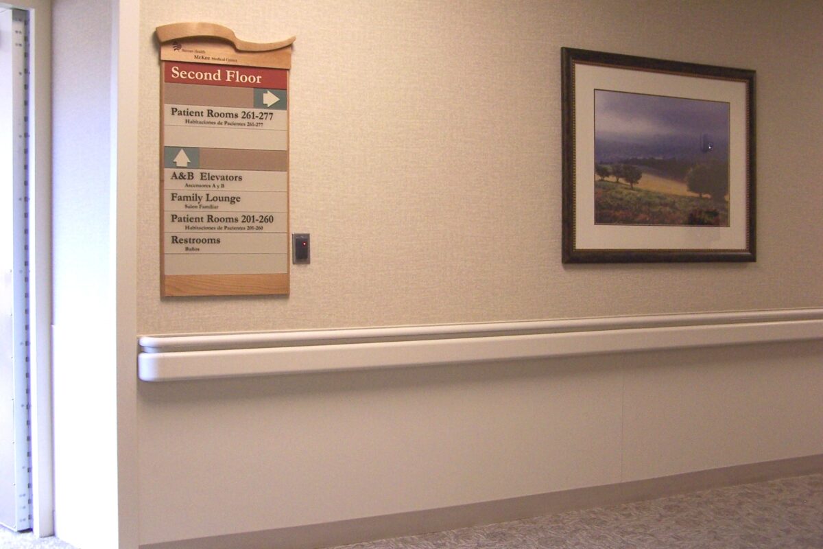 A beige hospital corridor with a wooden-framed directory on the wall indicating the second floor. The directory lists patient rooms 201-213 and other facilities. A framed nature photograph is on the adjacent wall. An open door is partially visible on the left.