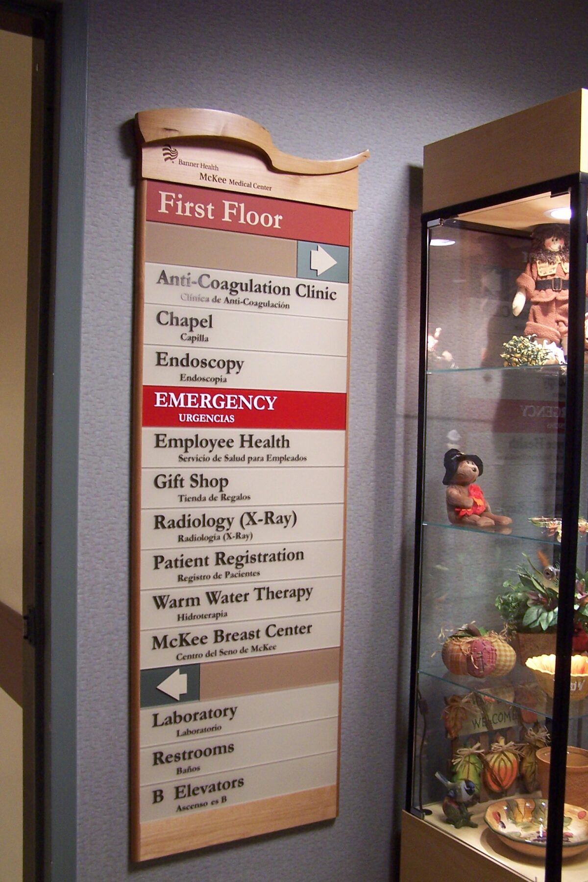 A directory sign on a wall lists locations on the first floor of a building, including the Anti-Coagulation Clinic, Chapel, Endoscopy, Emergency, Employee Health, Gift Shop, Radiology (X-Ray), Patient Registration, Warm Water Therapy, and McKee Breast Center.