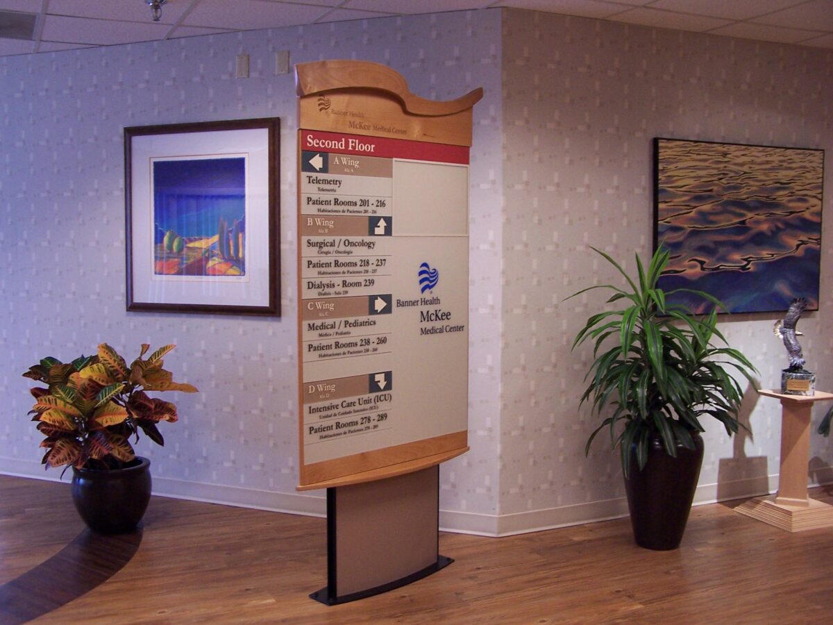 A hospital directory sign stands in a hallway, listing departments on the second floor. Two framed pictures hang on the wall behind it. Potted plants are positioned nearby, and the hallway has a wooden floor. .