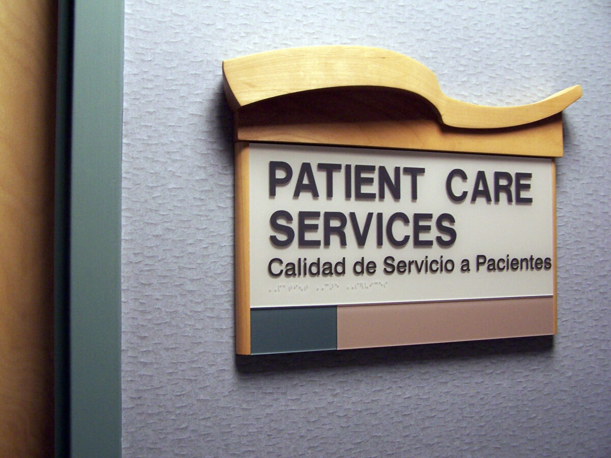A wall-mounted sign reads "PATIENT CARE SERVICES" with the Spanish translation "Calidad de Servicio a Pacientes" below it. The sign is bordered by a light wooden top section and colored panels at the bottom. The background wall is light gray.