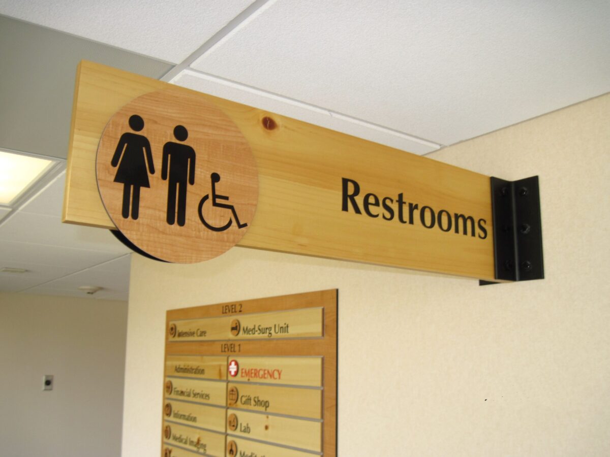 A wooden sign with a female and male restroom icon, a wheelchair accessibility symbol, and the word "Restrooms," is mounted on a beige wall. Below the sign is a directory listing various locations within the building.