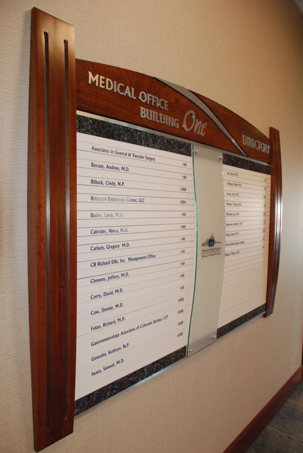 A directory sign inside a building labeled "Medical Office Building One" lists names and room numbers of various doctors. The sign has a wooden frame with a curved top and features a partial glass panel in the center.