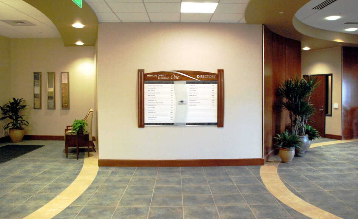 The image shows a spacious, well-lit interior of a building lobby with tiled flooring and neutral-colored walls. A directory board is mounted on the central wall, flanked by potted plants. A hallway is present on the right side, leading to other rooms or areas.