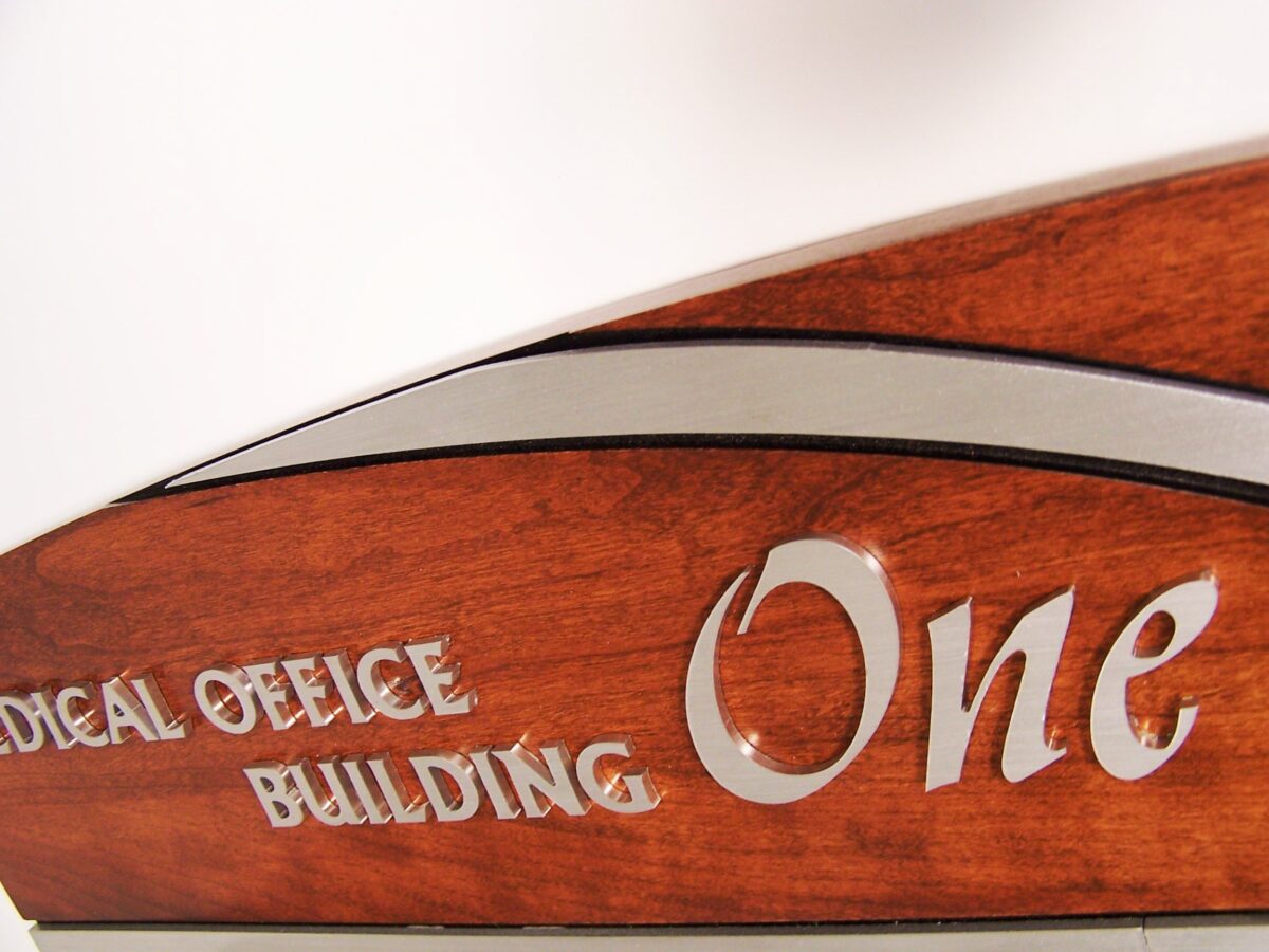 Close-up image of a wooden sign with metallic lettering that partially reads "Medical Office Building One." The focus is on the top edge of the sign, highlighting its curved design and polished wood finish.