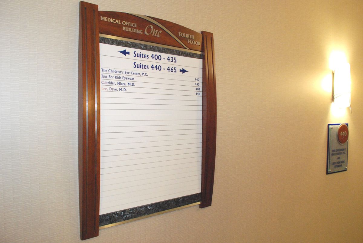 A mounted board with a wooden frame displays a list of events scheduled for the day. To the right, a wall-mounted directory sign can be seen. The board is titled "MEDICAL OFFICE" and indicates rooms and times for appointments. The setting appears to be in a corridor.