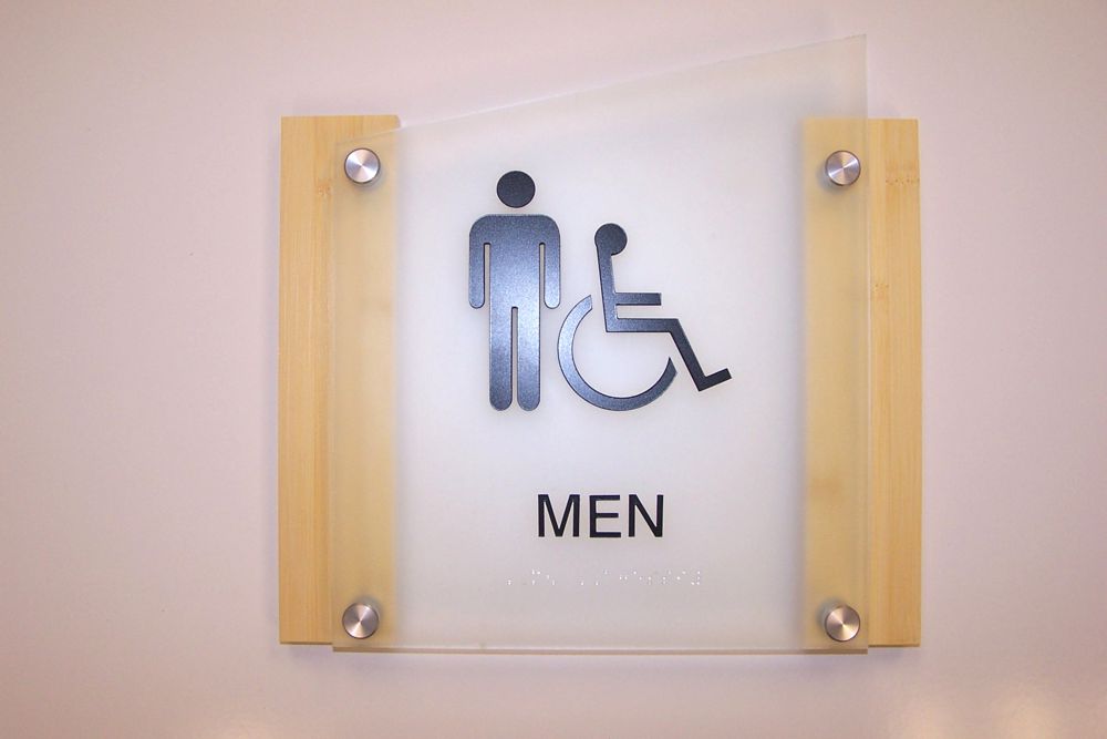 A restroom sign mounted on a white wall indicating a men's restroom. The sign features a male stick figure standing next to a wheelchair symbol, with the word "MEN" written beneath the icons.
