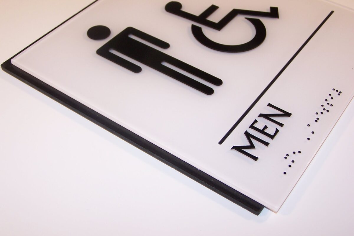 A white, rectangular restroom sign with black symbols indicating accessibility for men. The left side features a standard male figure icon next to a wheelchair accessibility icon. Below, the word "MEN" is written in black text, along with Braille.