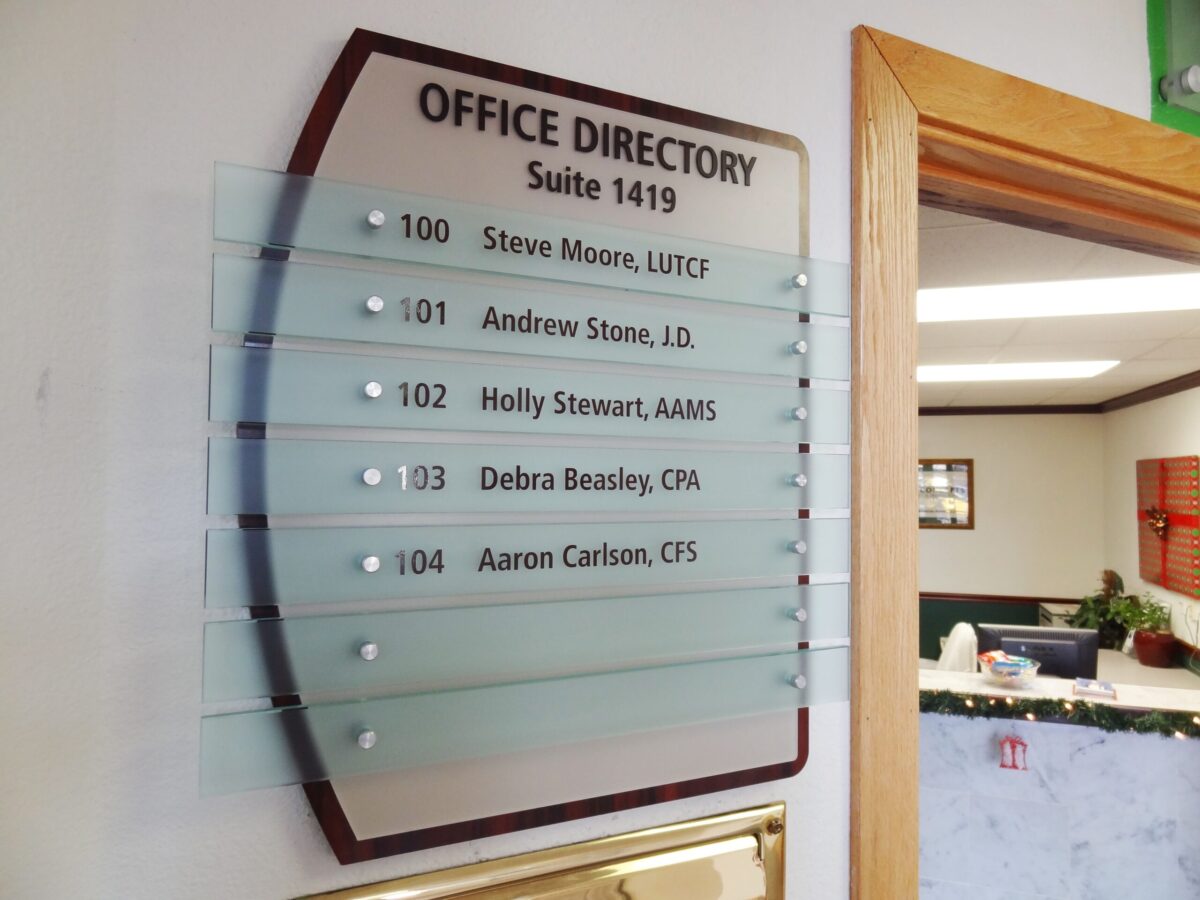 A glass office directory sign mounted on a beige wall lists suite 1419 occupants: 100 Steve Moore, LUTCF; 101 Andrew Stone, J.D.; 102 Holly Stewart, AAMS; 103 Debra Beasley, CPN; 104 Aaron Carlson, CFS. The entrance to a well-lit office space is visible to the right.