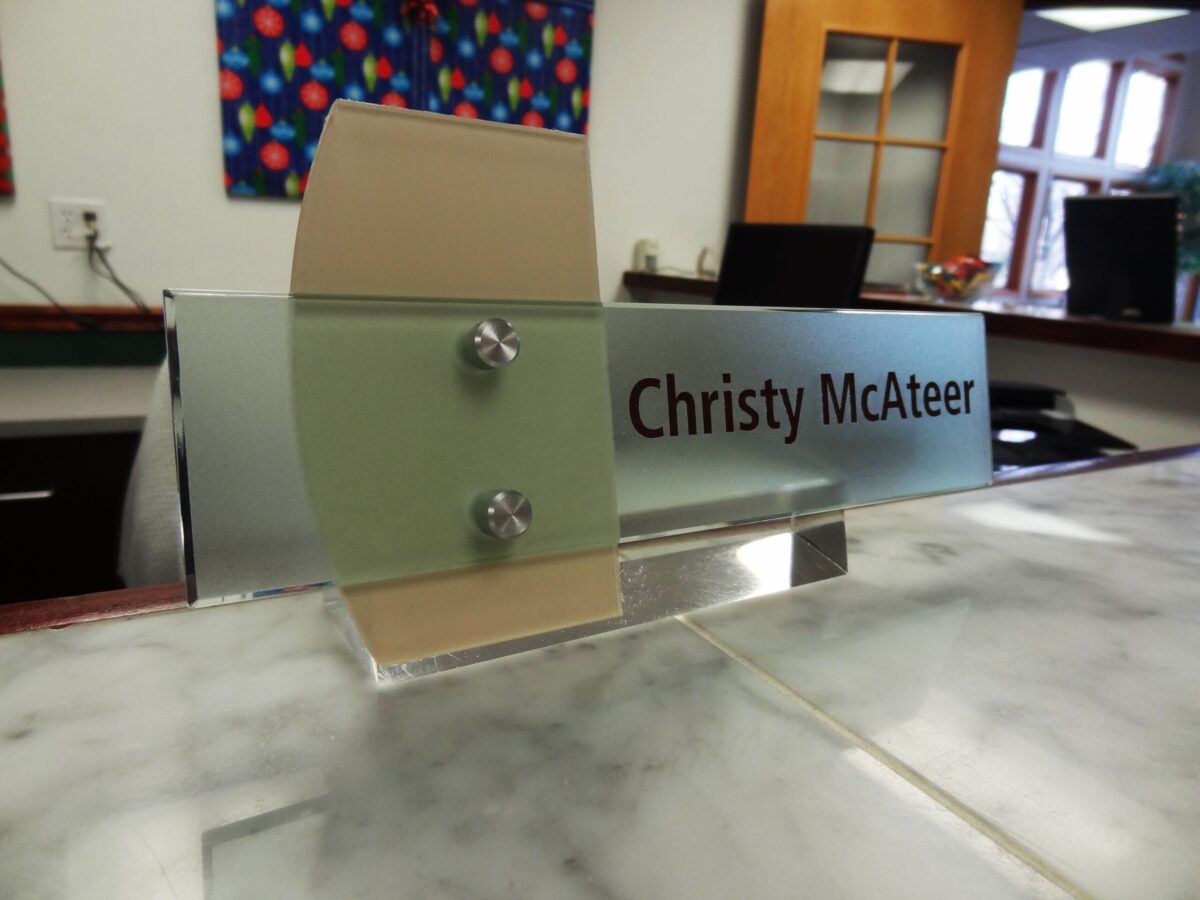 A nameplate on a marble counter reads "Christy McAteer." The background shows an office setting with a colorful painting, a wooden door with glass panes, and office chairs. Natural light is coming through windows.