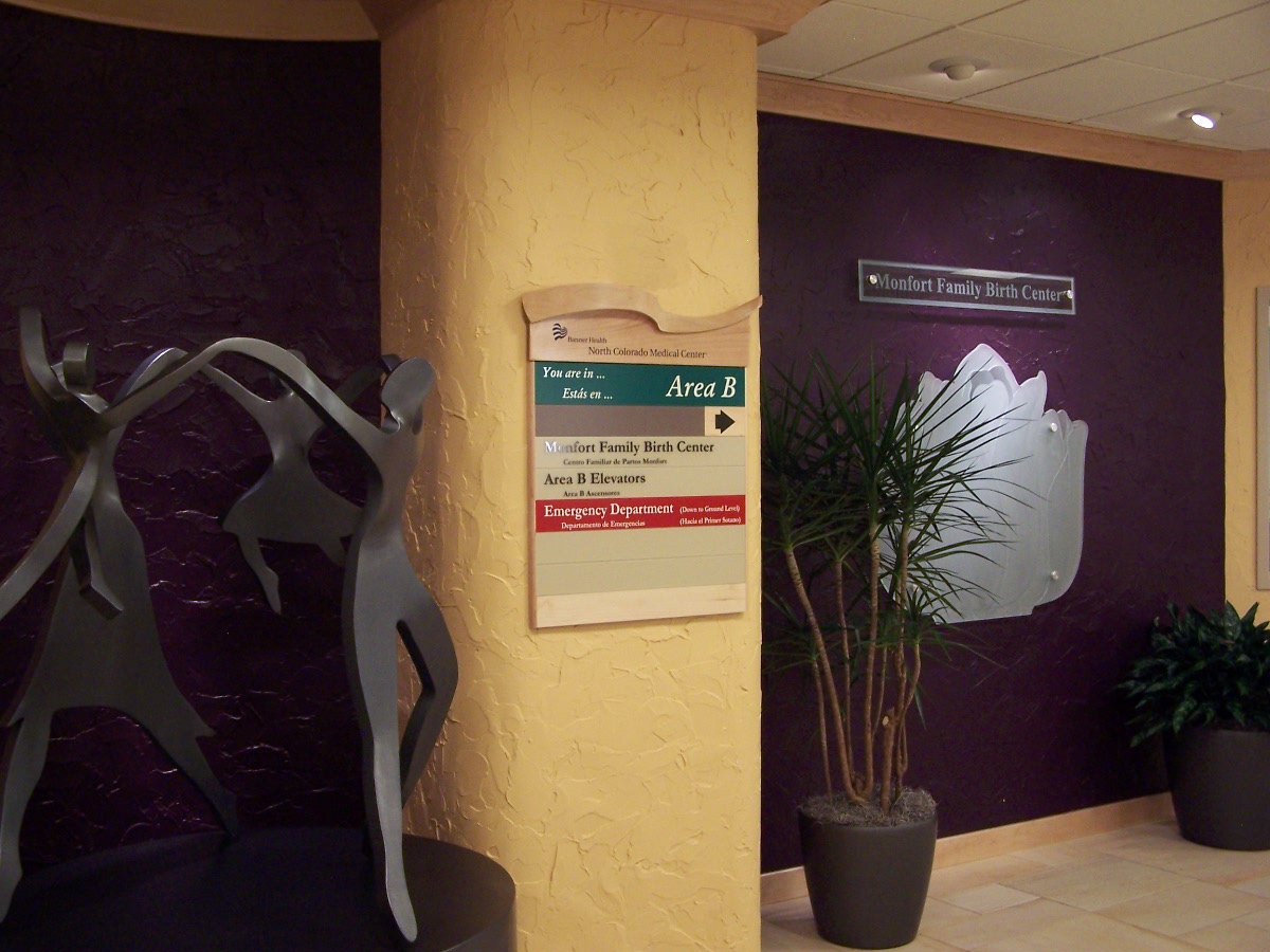 A hallway in a clinic or hospital with a sign directing to Area B, including family birth center and emergency department. There is a metallic sculpture of human figures to the left and potted plants on both sides. Walls have a textured and plain backdrop.
