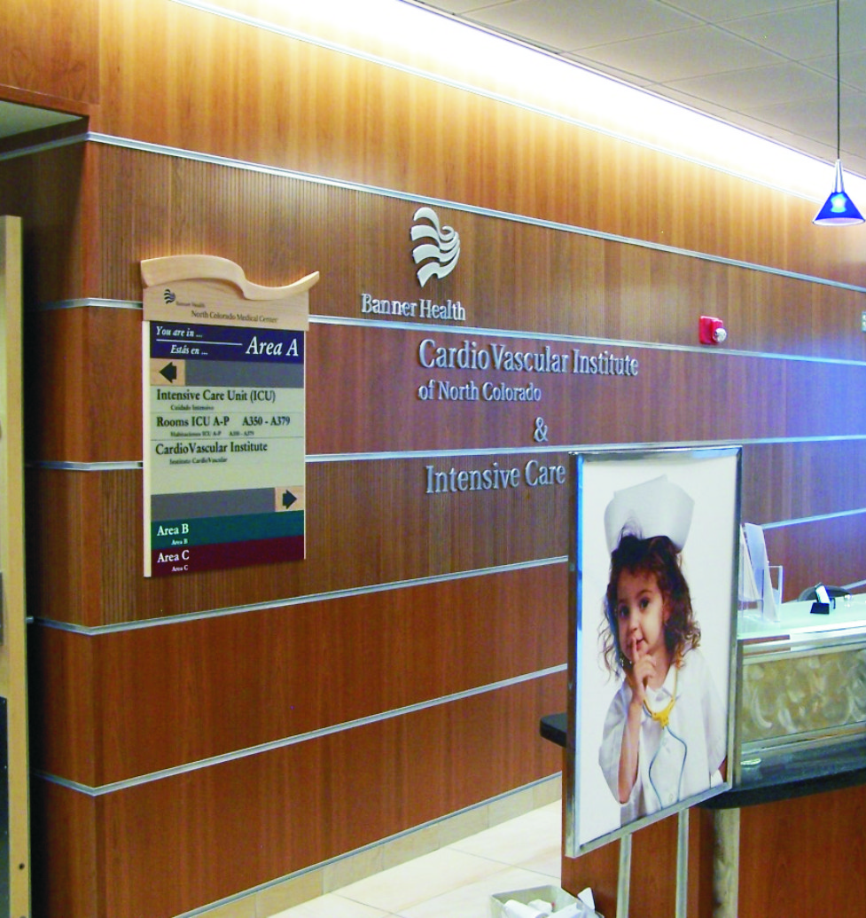A medical facility hallway with wooden panel walls and a sign that reads "Cardio Vascular Institute of North Colorado & Intensive Care." A signboard lists different areas, and a large poster of a child dressed as a nurse is displayed near a desk.