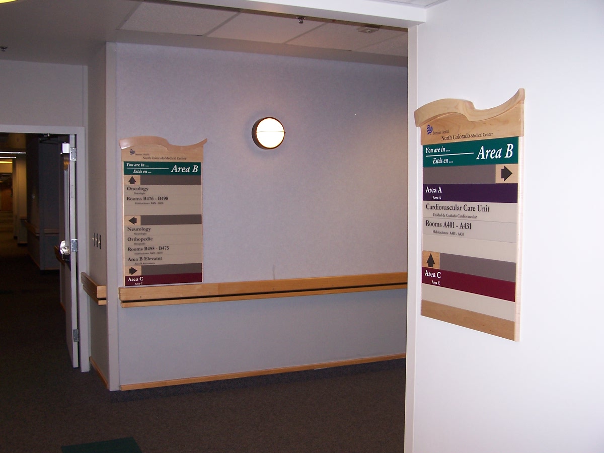 An interior hallway with two directory signs on the walls, labelled "Area B." Each sign lists multiple room numbers and departments in different colors. The walls are white and there is a circular light fixture in the middle of the wall.