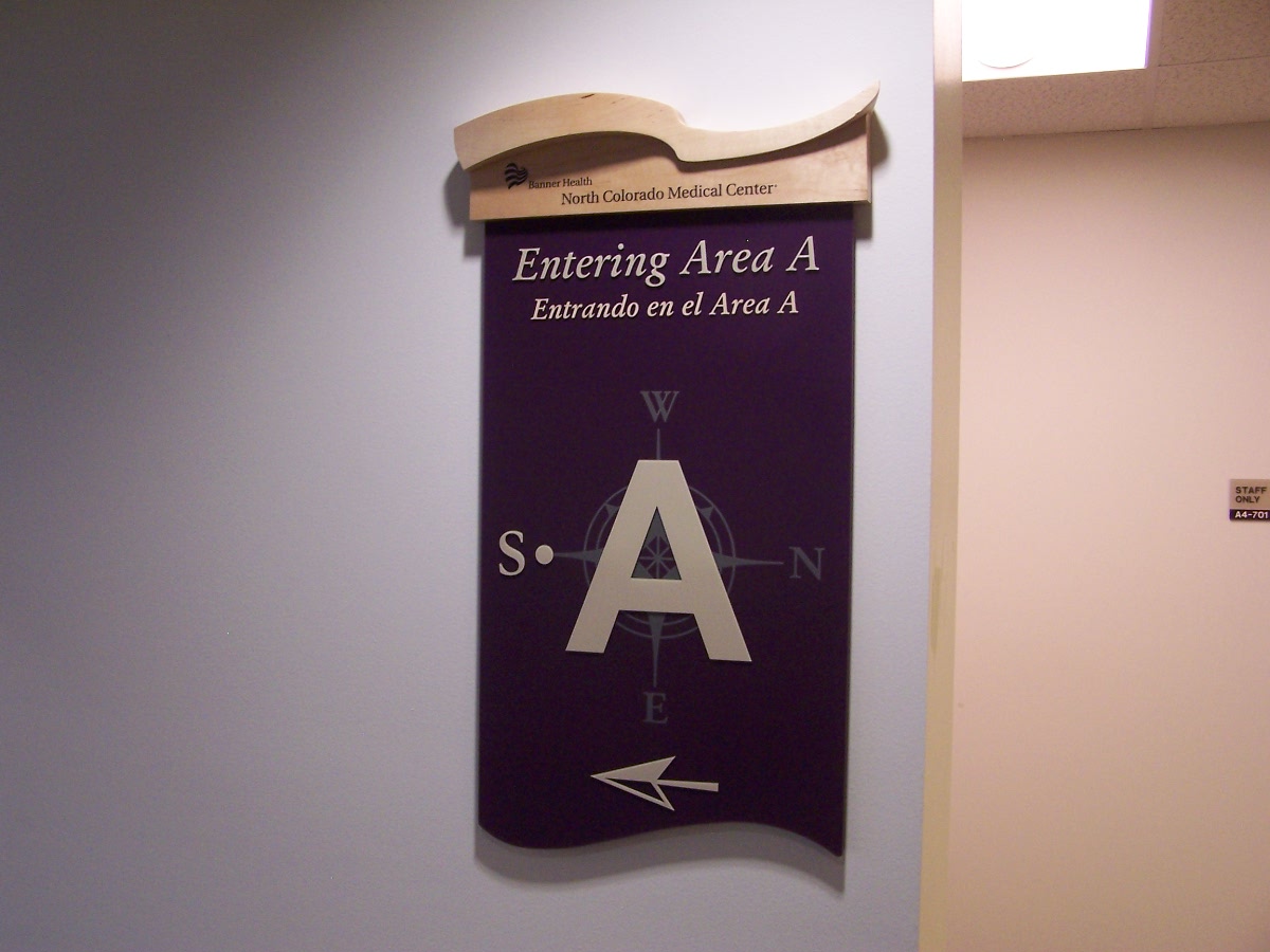 A sign on a wall reads "Entering Area A / Entras a el Area A" with a large letter "A" in the center and compass directions around it (N, E, S, W). An arrow pointing left is at the bottom of the sign. The top portion includes an arched decorative element.