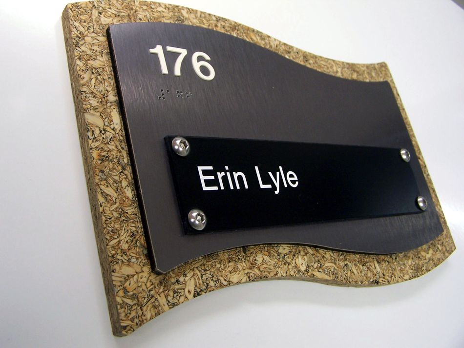 A wall-mounted, curved-edge nameplate with the number 176 at the top and 'Erin Lyle' written below on a black plaque. The border of the plaque has a textured, cork-like appearance.