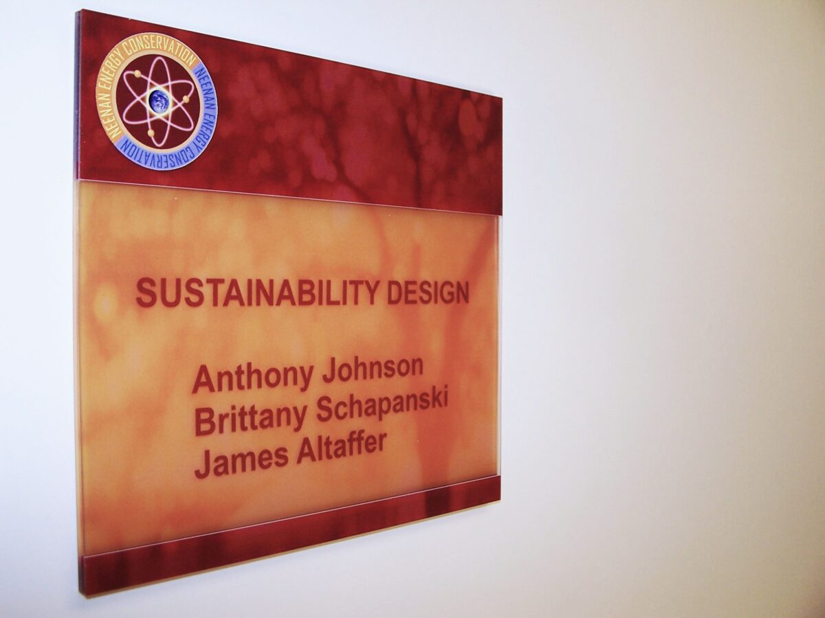 Plaque on a wall reading, "Sustainability Design" with names listed below: "Anthony Johnson, Brittany Schapanski, James Altaffer." The plaque has a red, marble-like background. The upper left corner features a logo with an atom design.