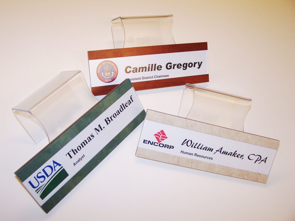 The image shows three desk nameplates. One is labeled "Camille Gregory" and has a logo on the left. Another reads "Thomas M. Broadleaf" with a USDA logo. The third nameplate is for "William Awaker, CPA" with an ENCORP logo. Each nameplate is on a clear stand.