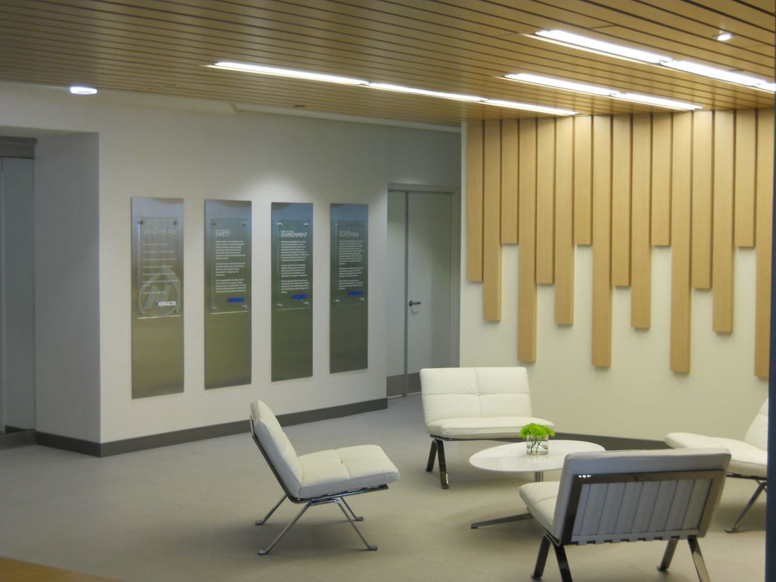 A modern office lobby with wooden paneling on the wall and a wooden ceiling. The lobby is furnished with white minimalist chairs and a small round white coffee table with a green decorative piece on it. Four framed pieces of art are displayed on the wall.