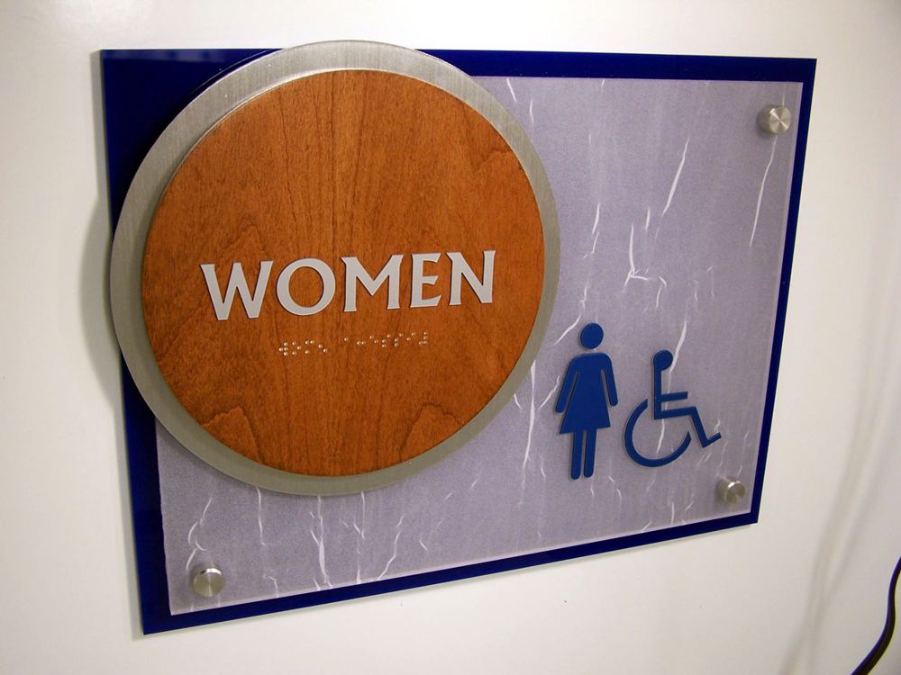 A restroom sign indicating a women's accessible restroom. The sign features a round, wooden plaque with "WOMEN" written in white text, and below it are icons of a female figure and a wheelchair symbol, both in blue.