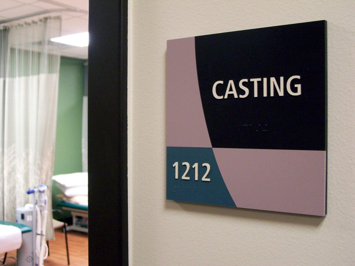 A sign on a wall reads "CASTING 1212." Beyond the doorway, two medical examination tables are visible in a clinical room setting with light green walls and a curtain divider.