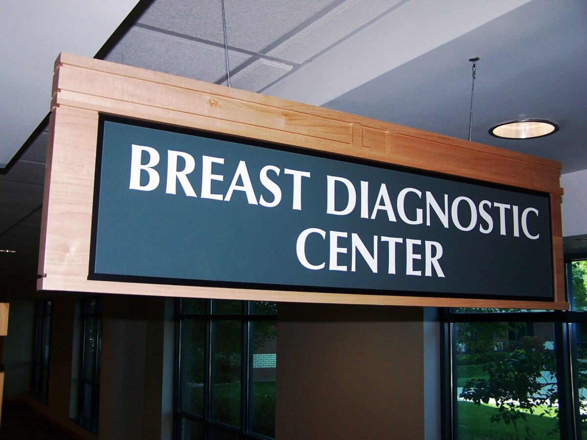 A rectangular wooden sign with a dark blue background and white text hangs from a ceiling, reading "BREAST DIAGNOSTIC CENTER." The sign is located indoors, with windows and fluorescent lights visible in the background.