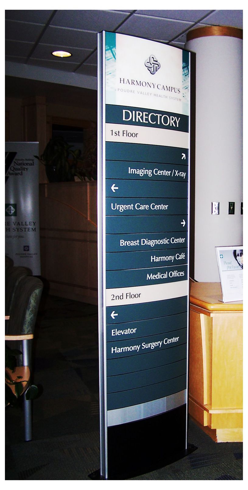 A standing directory sign in a medical facility lists locations for various departments: Imaging Center, Urgent Care Center, Breast Diagnostic Center, Harmony Café, Medical Offices, Elevator, and Harmony Surgery Center, with arrows indicating directions.