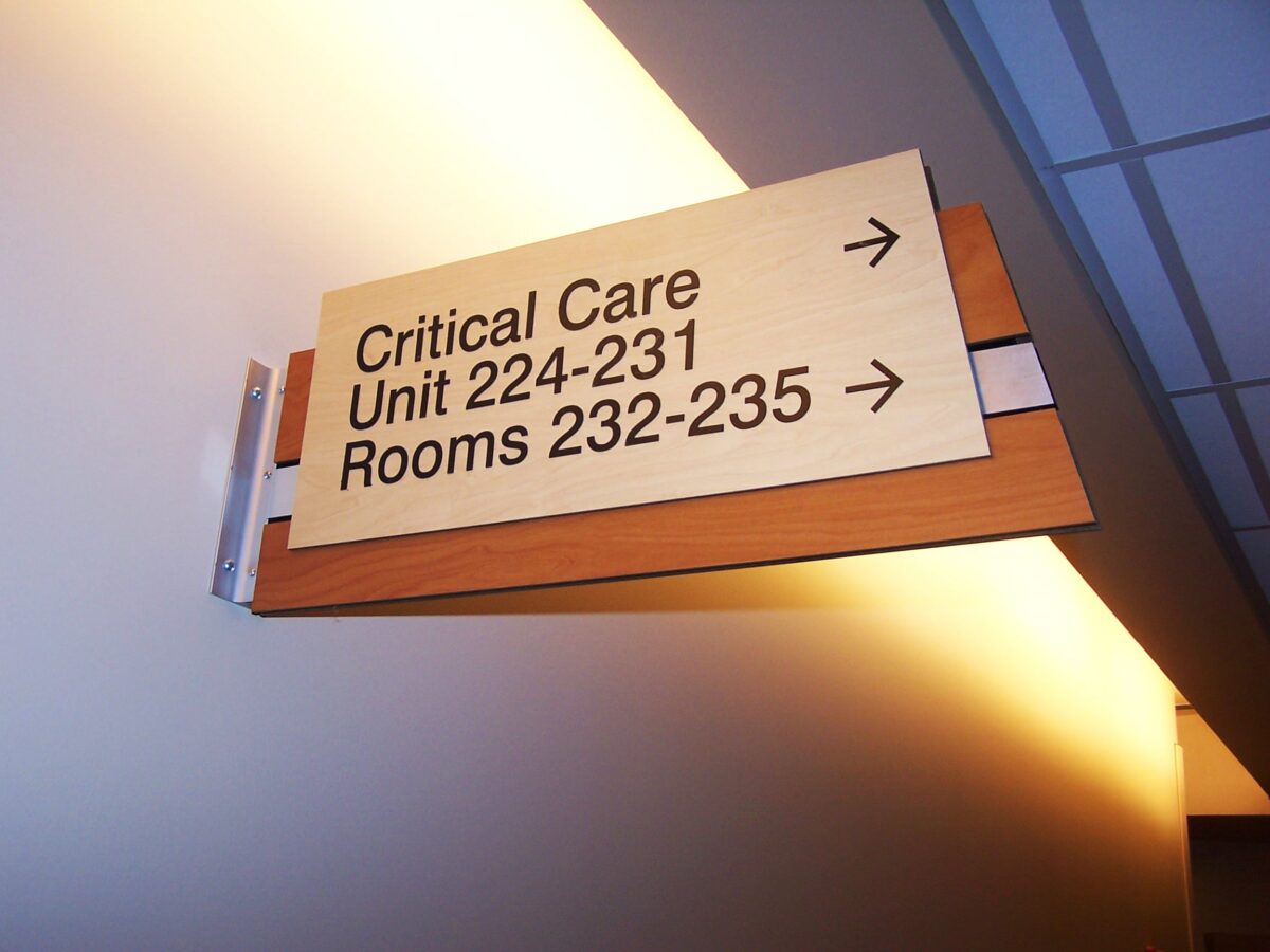 A hallway sign indicating directions to "Critical Care Unit 224-231" and "Rooms 232-235" with arrows pointing right. The sign is mounted on a wooden bracket on a lightly colored wall, and the ceiling features recessed lighting and ceiling tiles.
