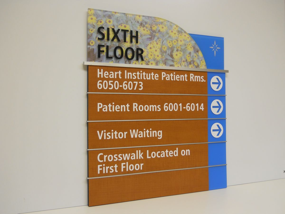 A wall-mounted sign indicating directions on the sixth floor of a building. It lists Heart Institute Patient Rooms 6050-6073, Patient Rooms 6001-6014, Visitor Waiting, and mentions a crosswalk located on the first floor, with arrows pointing right.