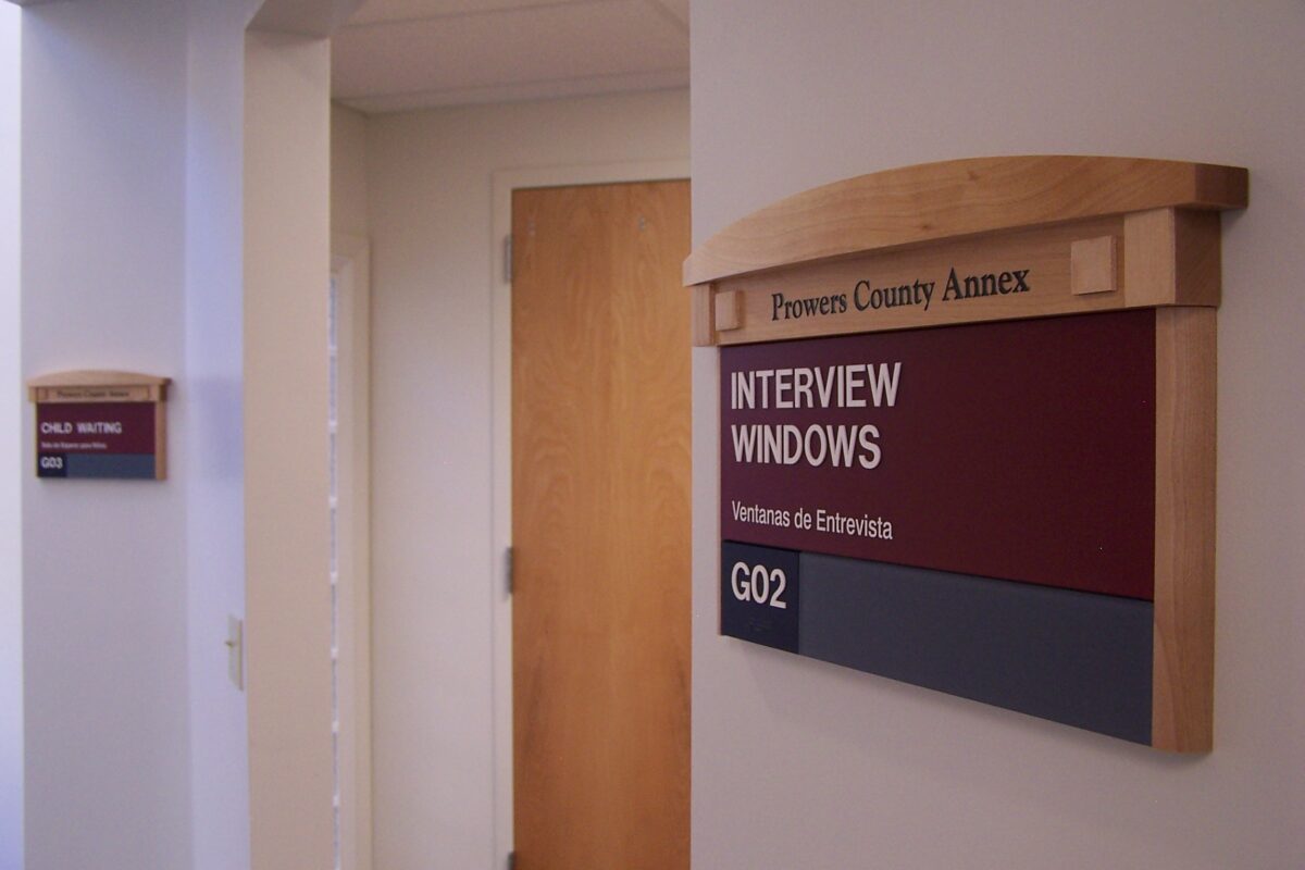 A hallway in a building with a sign on the wall reading "Powers County Annex" and "Interview Windows" with room number "G02." A smaller sign indicating "Public Restroom" is also visible in the background near a door.