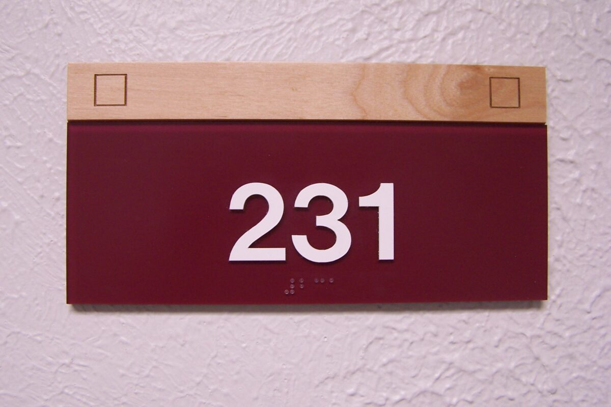 A rectangular room number sign reading "231" is mounted on a textured white wall. The sign has a dark red background with white numbers and a wooden strip at the top. The sign also has two square indents on the wooden strip.