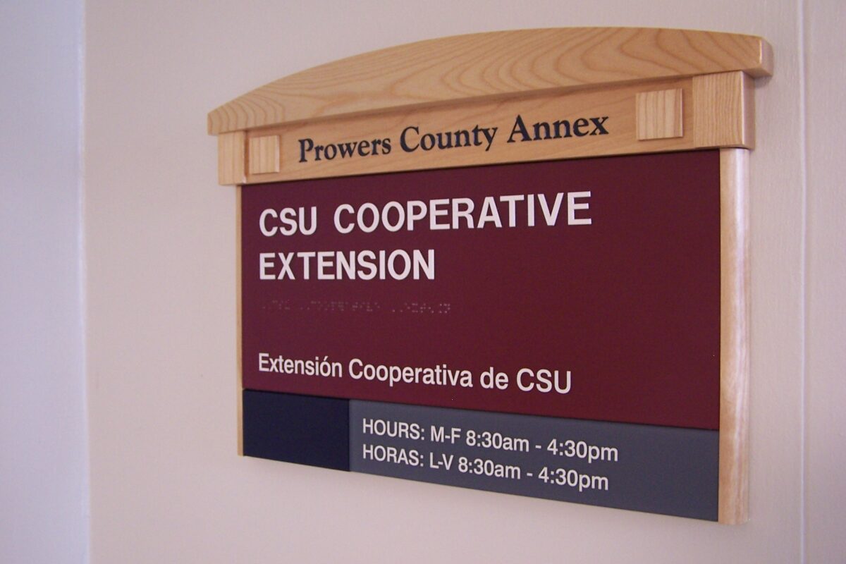 A sign on a wall reads "Prowers County Annex" at the top. Below, it says "CSU Cooperative Extension" and "Extensión Cooperativa de CSU." The hours of operation are listed as Monday to Friday, 8:30 am - 4:30 pm in both English and Spanish.