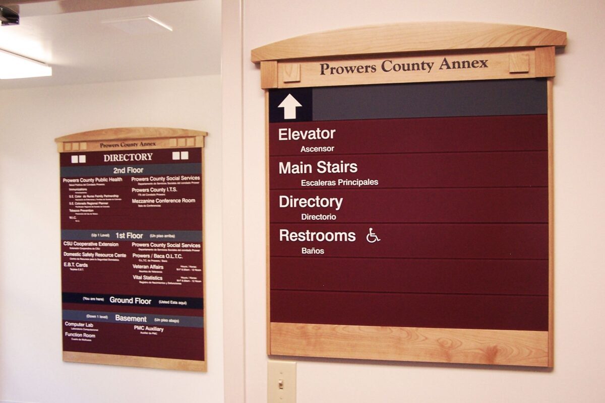 Two signs in a building hallway provide directions. The larger sign has labels in white text for elevator, stairs, directory, and restrooms. The smaller sign lists detailed information for services on different floors. The top of both signs reads "Prowers County Annex.