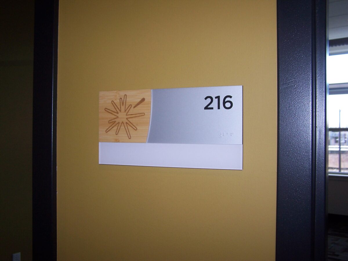 A room number sign on a yellow wall displays the number 216 in black text on a silver background. Beside the number is a design of abstract lines on a light brown background.