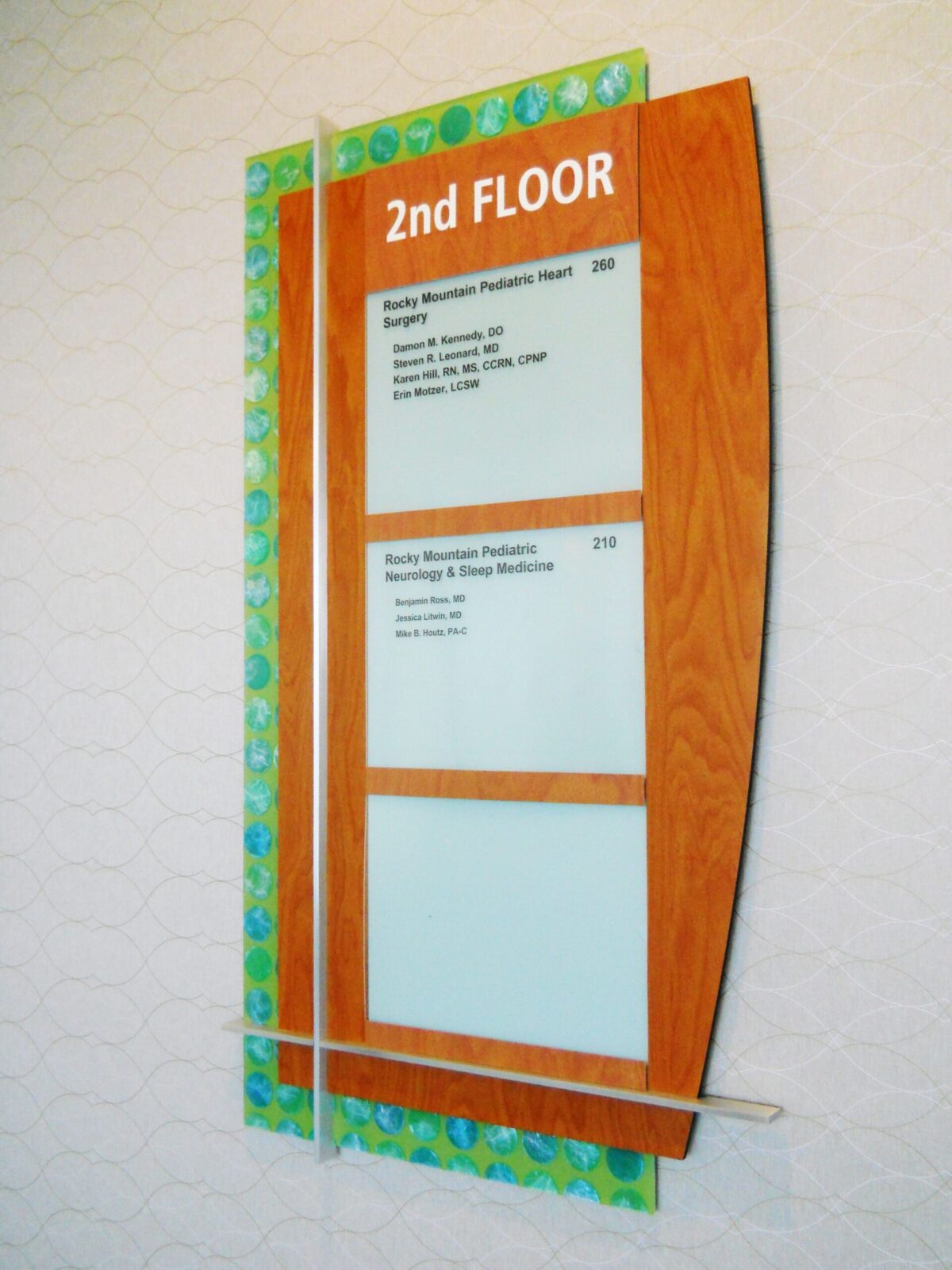 A directory sign for the 2nd floor on a wall. The sign includes a list of departments and room numbers in brown and white lettering. The sign has a wood-textured frame with a decorative blue-green border. The background wall has a hexagonal pattern.