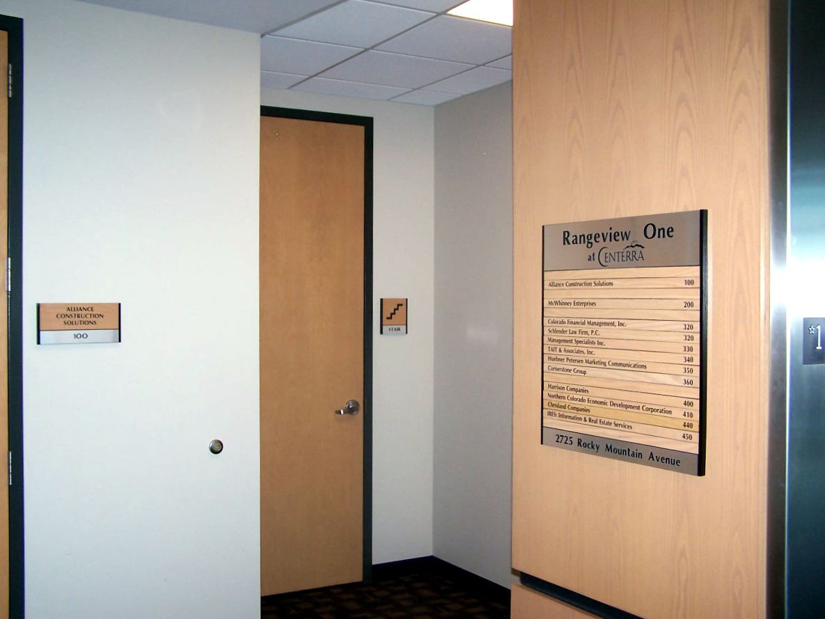 A hallway with off-white walls and a light wooden door. On the wall, there is a large directory sign listing various rooms and their locations. A smaller sign is mounted on the adjacent wall to the door, displaying additional information. The lighting is bright and even.