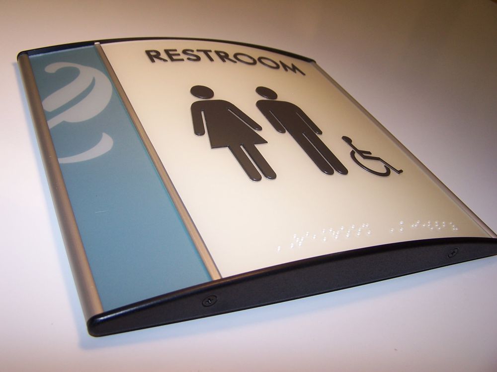 A restroom sign featuring icons for men, women, and wheelchair accessibility. The sign has a blue and white color scheme with a curved, modern design.