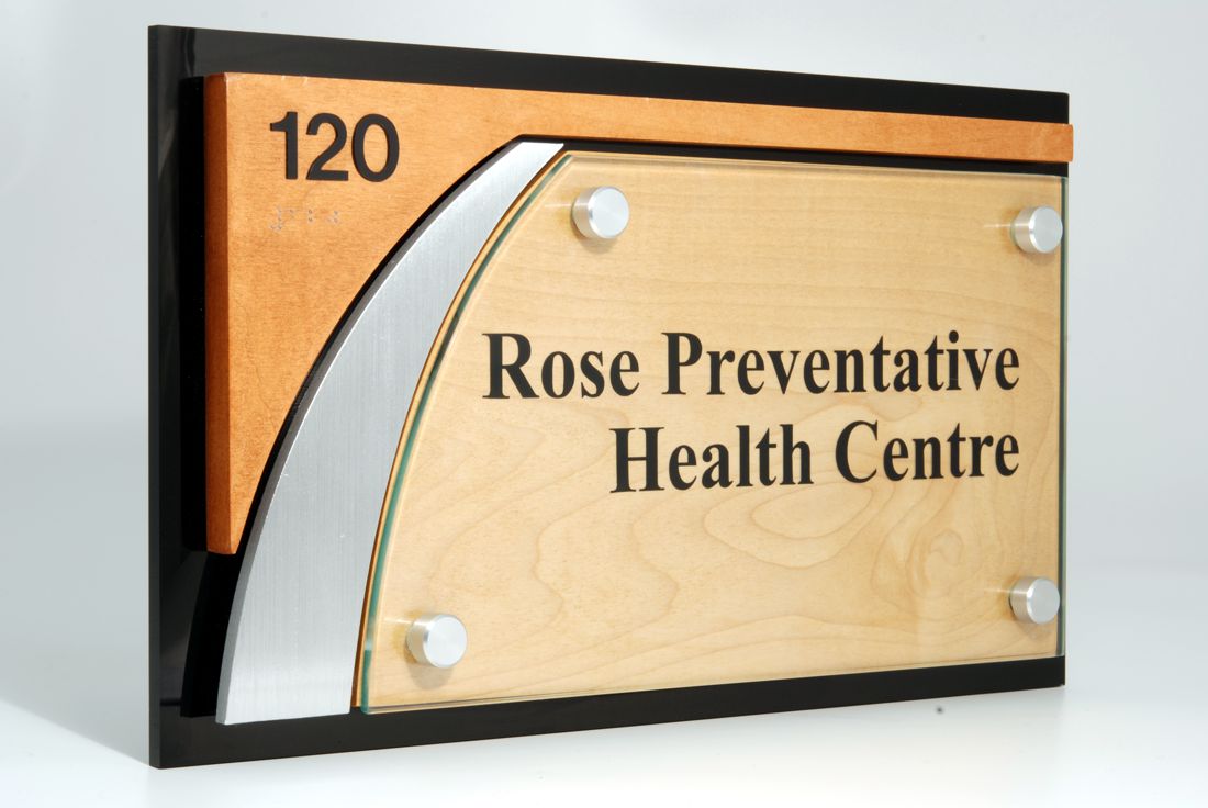 A wall sign with the number "120" in the upper left corner. The sign reads "Rose Preventative Health Centre" written on a light wood background, bordered by a black frame with a modern metal accent on the left-hand side.
