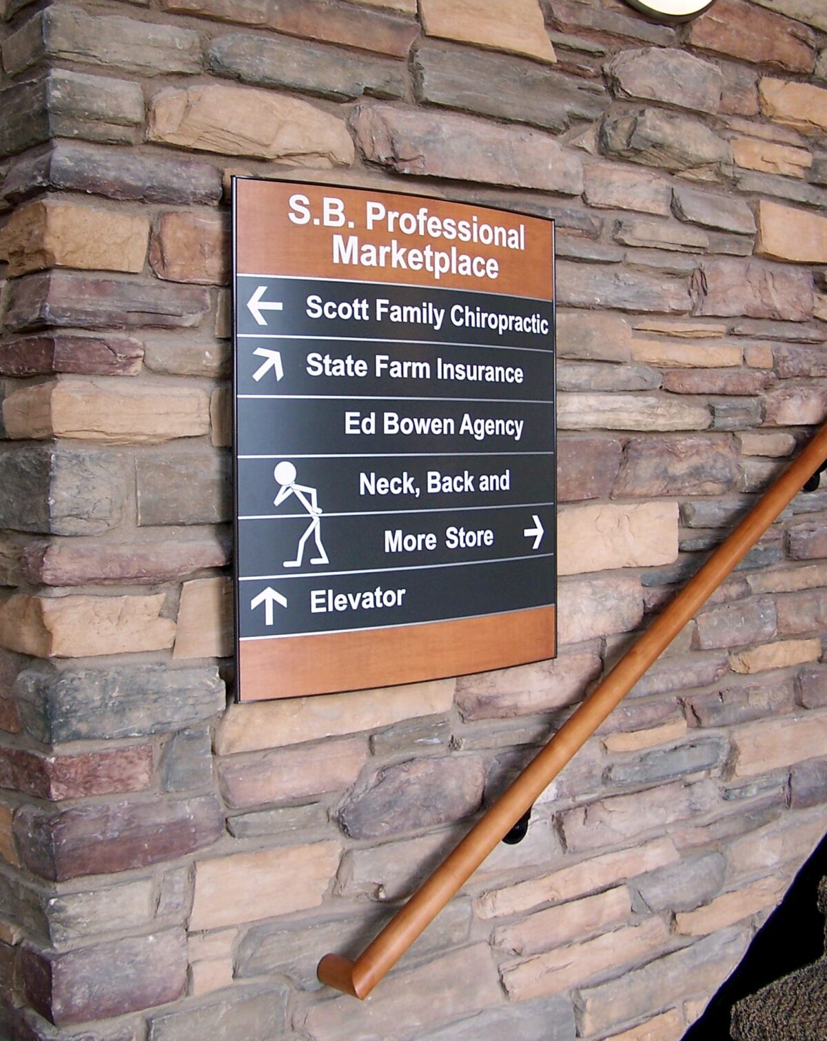 A directory sign on a stone wall indicates businesses in the S.B. Professional Marketplace. It lists: Scott Family Chiropractic, State Farm Insurance, Ed Bowen Agency, Neck, Back and More Store, with directions and an arrow pointing towards the elevator.