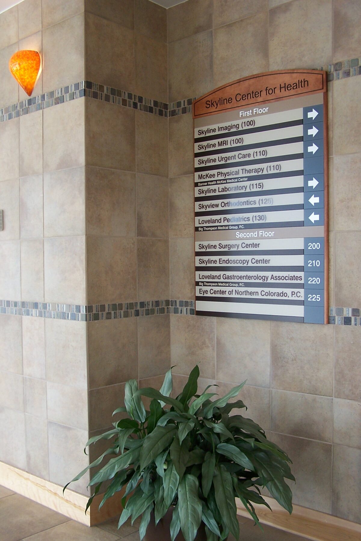 A directory sign mounted on a beige, tiled wall in a building with directions to various departments. A green plant in a pot sits at the base of the wall. An orange sconce light is visible above the sign. The area appears clean and well-maintained.