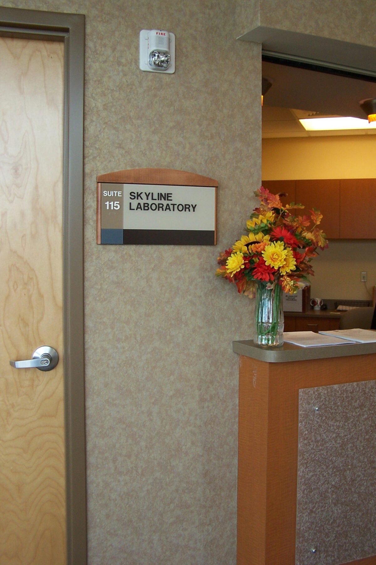 A laboratory door labeled "Suite 115 Skyline Laboratory" next to a beige reception counter with a bouquet of colorful flowers in a glass vase. The lab appears to be inside a building with beige textured walls and an open door, leading to a room with cabinets and bright lighting.
